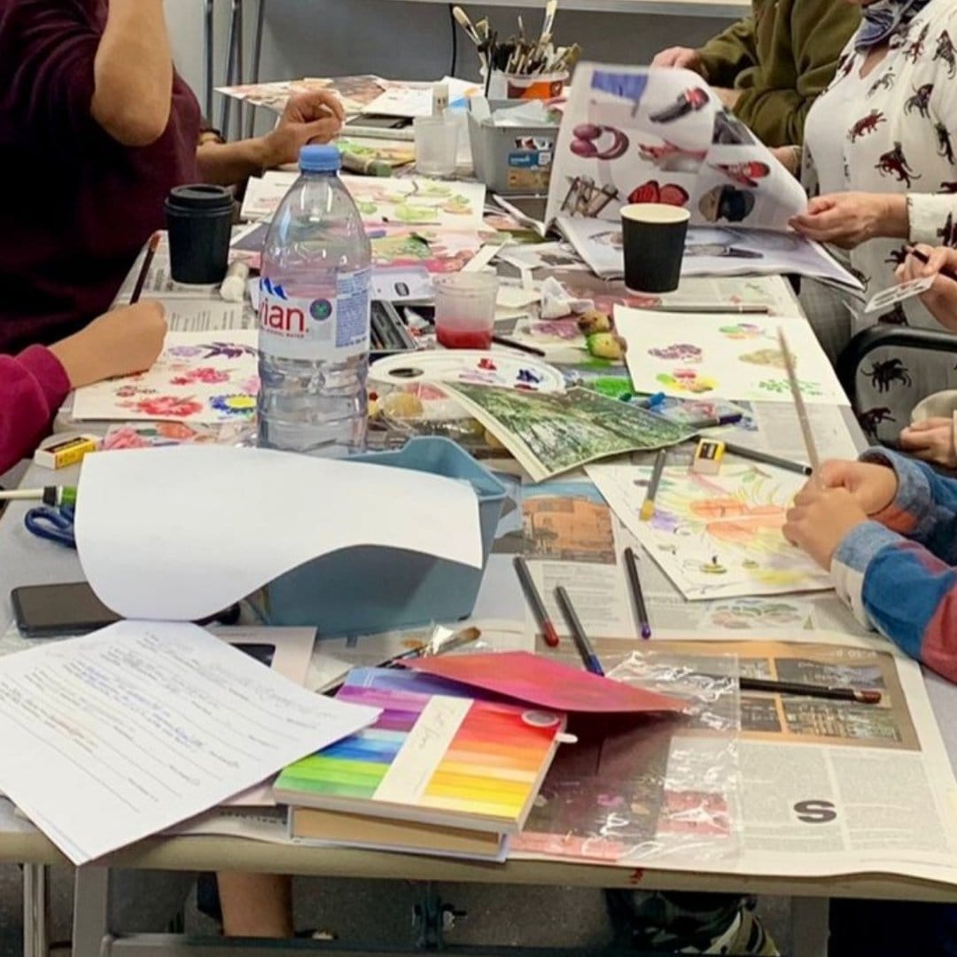 A workshop taking place, visible is a table covered in paints and craft materials and four peoples hands making things.