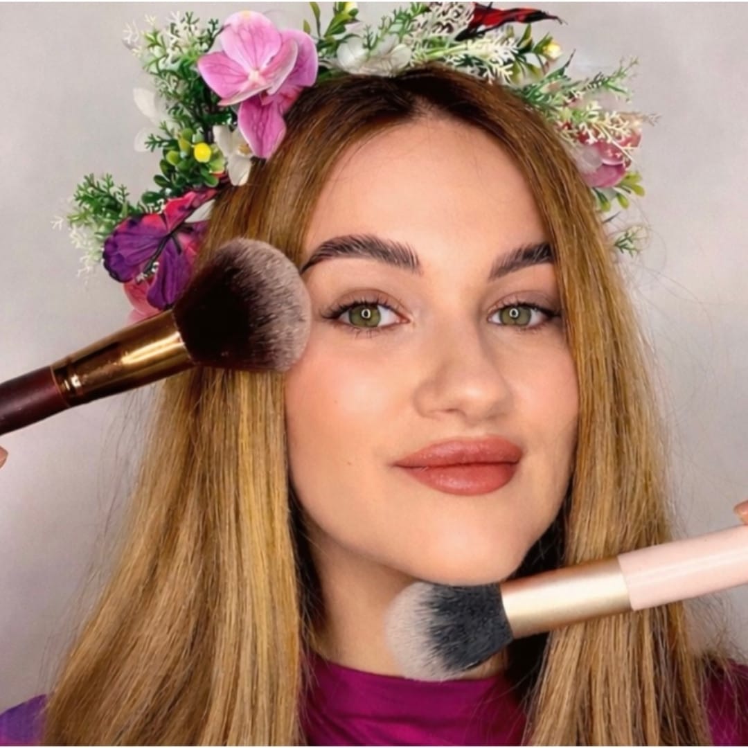 An image of a young woman with a flower crown on her head and two make up brushes touching her face