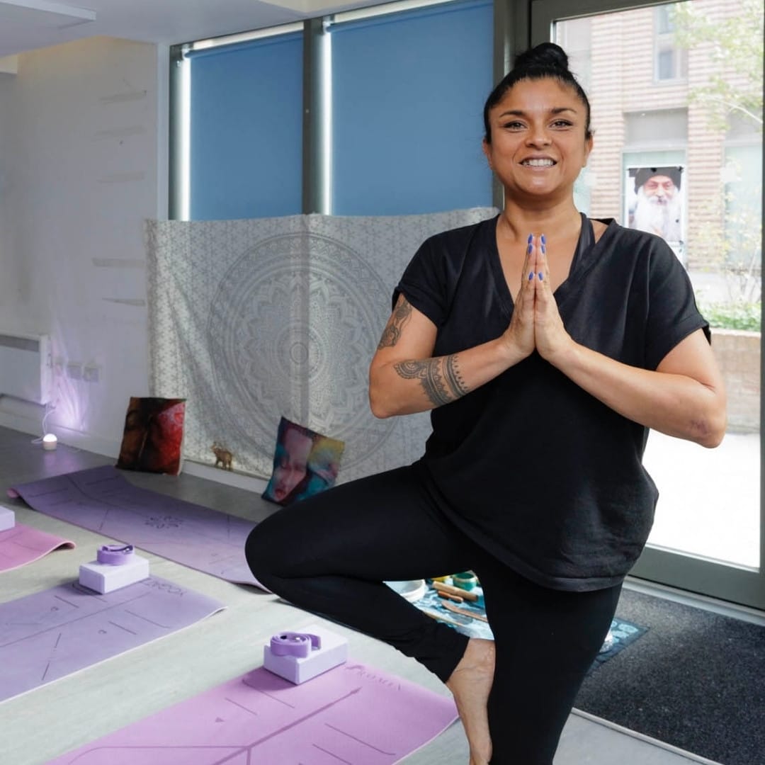 A woman in black leggings and tshirt does a yoga pose and smiles at the camera